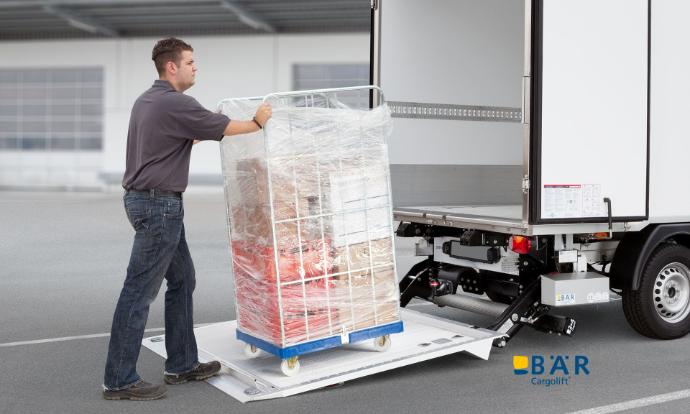 Un homme conduit un conteneur roulant sur un hayon élévateur BAR ouvert au sol.