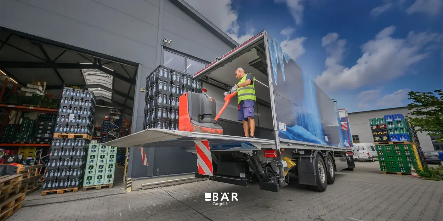 Un homme décharge une palette de boissons à l'aide d'un transpalette, via un hayon Bar Cargolift monté à l'arrière de la semi-remorque d'un ensemble articulé